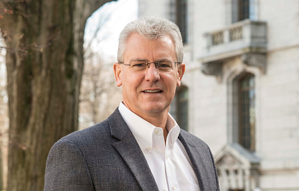 Johnson stands outdoors in front of a tall tree. In the background is a multistory white building with multiple windows and balconies.