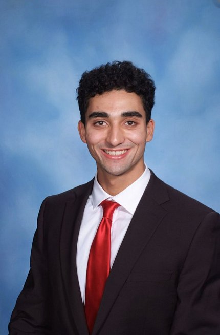Puthan, wearing a black suit, white shirt and red tie, against a blue background.