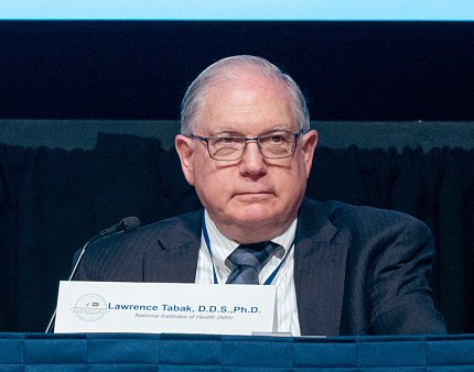 Tabak sits, with name placard, at a conference table.