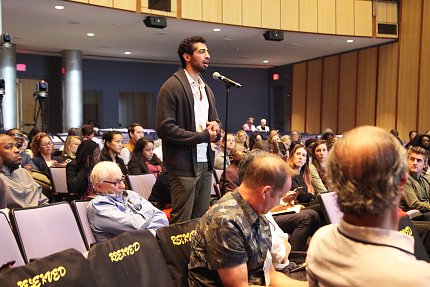 Man in brown cardigan stands to speak into a microphone with audience members seated around him