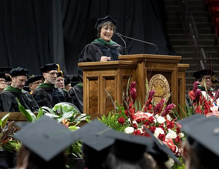 Bertagnolli in black cap and gown stands at podium as graduates wearing caps look on.