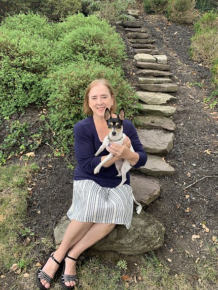 Ostrander seated on the bottom step of a rock path. She is smiling and holding a small black and white dog.