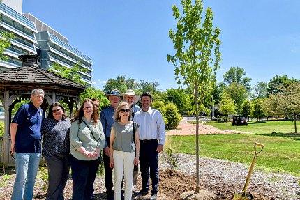 Several men and women outside on grounds of lab building smile beside newly planted tree.