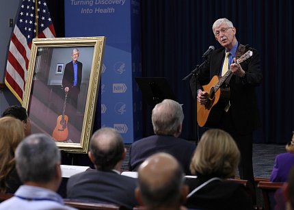 Collins plays guitar and sings to crowd, with portrait in the background