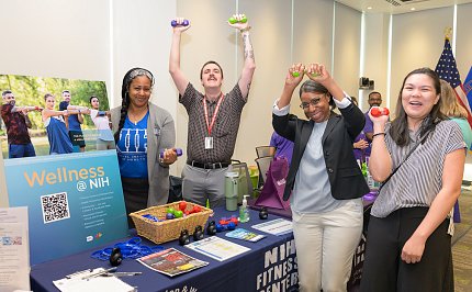 Several people stand by table, one woman squeezing a ball and a man holding up dumbells