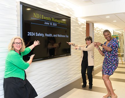 NIH director and two event organizers smile while pointing to Wellness Day sign on large TV screen.