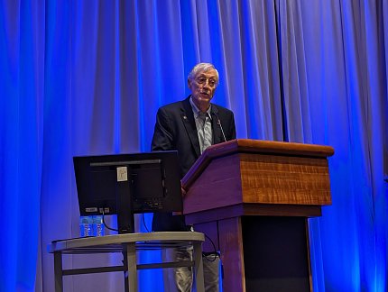 Mather speaks at a lectern, against the backdrop of a curtain lit by blue lights.