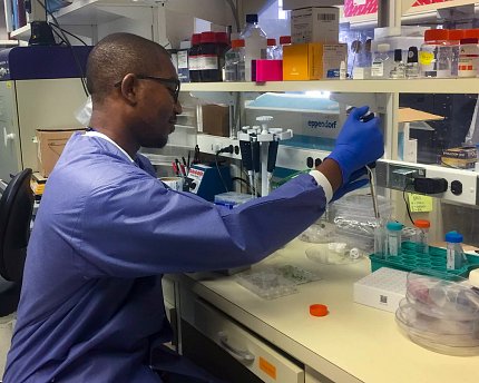 Nelson, wearing a blue lab coat and gloves, uses a pipette at a lab bench.
