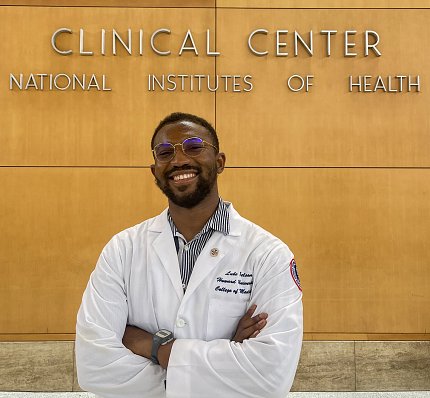 Nelson, wearing a white lab coat, poses in front of a light-colored wood wall. 
