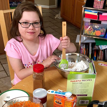 Abby, wearing glasses and a pink shirt, sits at a kitchen table, stirring a bowl of baking ingredients. The tabletop is covered in baking supplies.