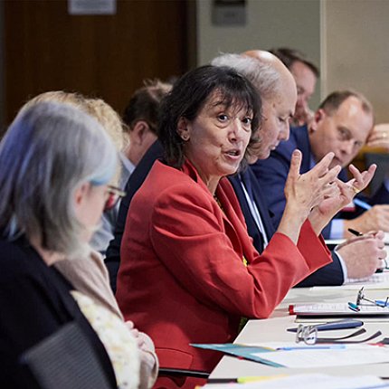 Bertagnolli, in red suit, holding up hands, talks at conference table
