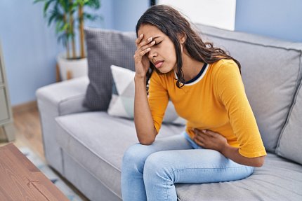Teenage girl seated on a couch rubs her stomach and makes a pained expression.