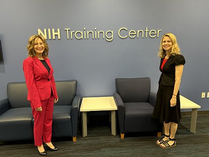 Two smiling women stand next to a wall with sign: "NIH Training Center" and table, chairs