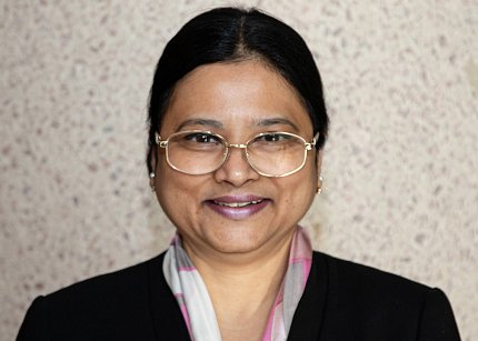 Desai, wearing large glasses and a black jacket, smiles as she stands against a light-colored wall.