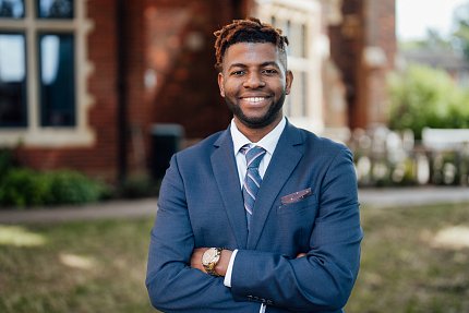 Tunyi smiles with his arms folded, standing outdoors with a brick building in the background.