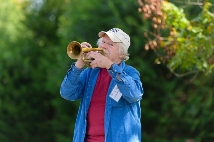 Seifried, wearing a white hat and denim shirt, jauntily plays a trumpet.