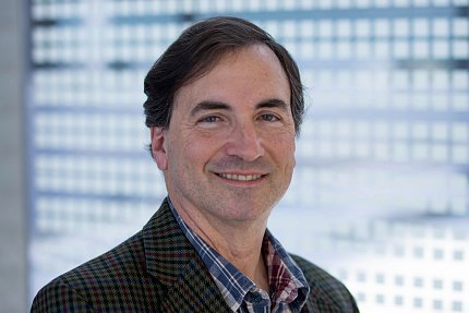 Headshot of Basser, wearing a brown jacket, silhouetted against a light blue background.