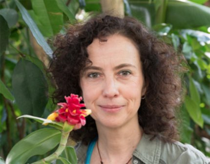 Vandebroek smiles softly in front of a leafy green plant. A red flower extends close to the right side of her face.