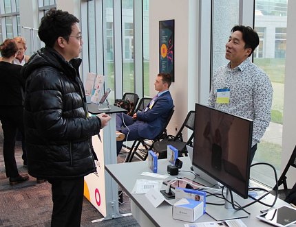 Two people stand on either side of table in hallway, talking, with computer monitor on table