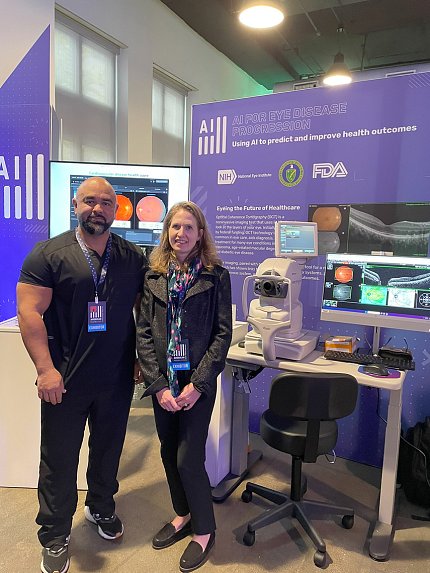 Arango and Goetz pose with their exhibit: a white ophthalmologic device and computer monitor on a desk, with a large purple poster in the background. 