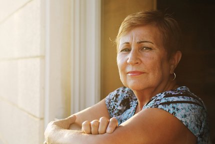 A woman sits facing the camera with her arms folded. A window that is mostly out of frame illuminates the front of her body.