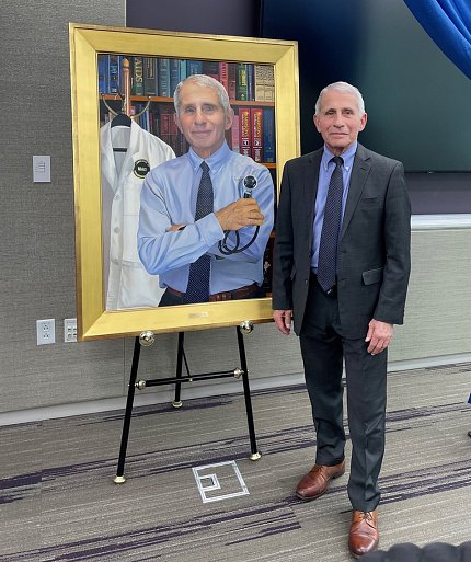 Fauci stands next to a framed painting of himself that shows him holding a stethoscope, in front of book case, with his lab coat hanging on a coat rack.
