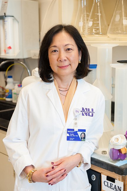Zee, wearing a white lab coat, leans against a counter holding laboratory equipment. 