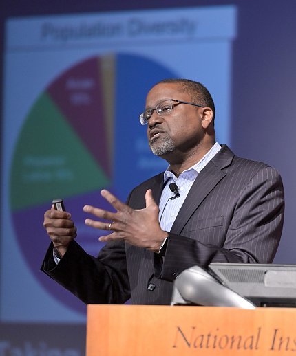 Gibbons speaks at podium with colorful pie chart on screen behind him.