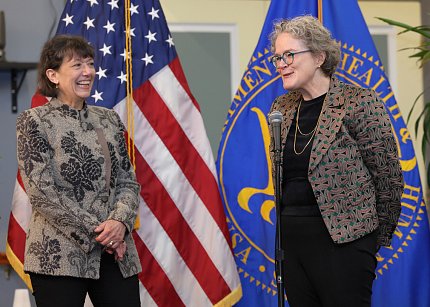 Bertagnolli laughs as Marrazzo delivers a speech. Behind them are the large American and Health and Human Services flags.