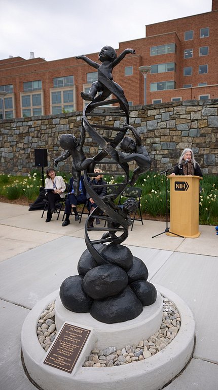 Statue of DNA double helix with four infants ascending it. Several seated individuals and one at a podium look on in the background.