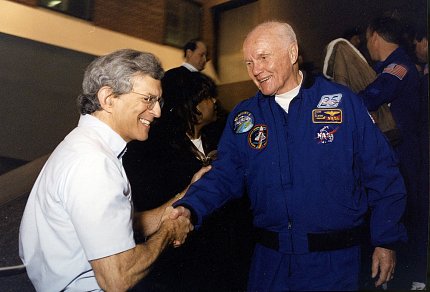 Smiling man reaches out to shake hands with man in astronaut uniform.