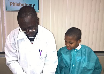 Phlebotomist Michael Guyah does a demonstration in the Clinical Center as Petty, then a young child, dressed in scrubs, looks on.