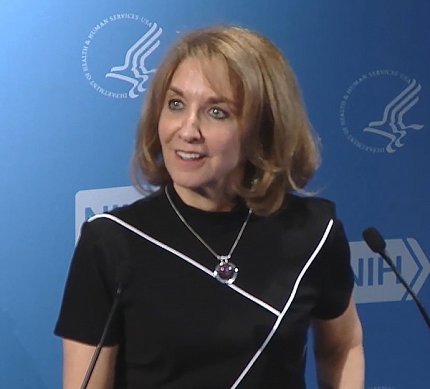 Kaplan, wearing a black outfit with diagonal white stripes, addresses an out-of-frame audience. A blue backdrop bearing the NIH and HHS logos fills the background.