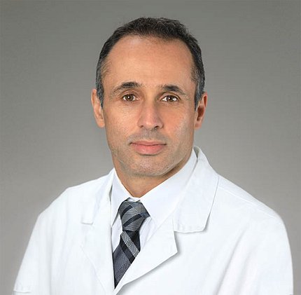 Zaghloul, wearing a shirt and tie and white coat, against a light gray background.