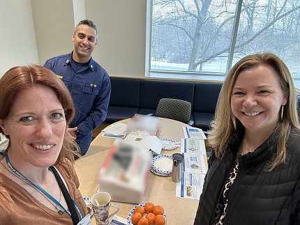 Three individuals pose for a selfie around a table filled with coffee and breakfast items.