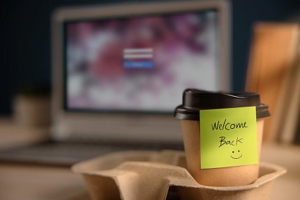 A lone coffee cup sits in a cardboard carrier. A sticky note on the cup bears a smiley face and the message: "Welcome back!"