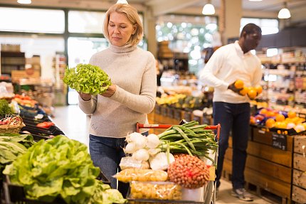A woman examines a leafy green vegetable in a grocery store. More fresh produce is piled in her shopping cart. In the background, a man is holding several oranges.