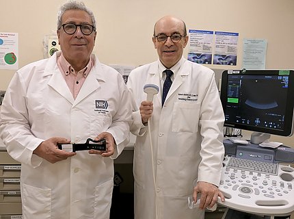Gandjbakhche and Romero pose for a photo. Both wear white coats; Gandjbakhche holds the biosensor and Romero holds an ultrasound transducer.