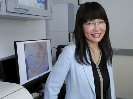 Image of Suh, wearing glasses and a blue jacket with one hand resting on a desk. Behind her is a computer screen with a scientific image.