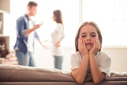 A child has her hands on her chin while adults fight in the background