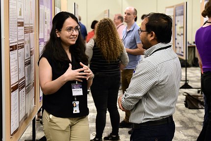 Fouladi gestures as she explains her research to an onlooker. A line of posters stretches out behind her, with crowds of other presenters and attendees. 