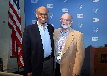 Goldstein and Nath stand in front of a light blue backdrop patterned with the NIH and HHS logos.