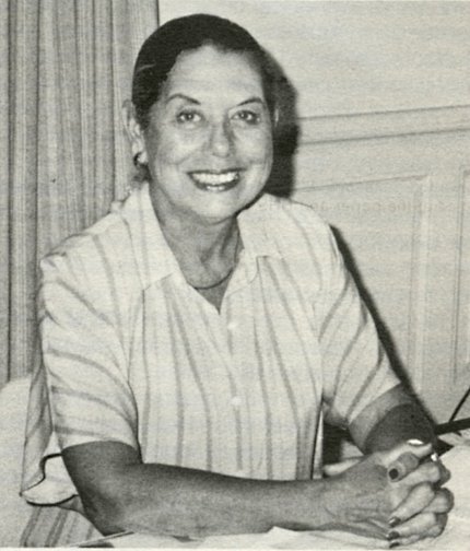 Black &amp; white photo of a smiling Bel Ceja, sitting hands clasped at her desk