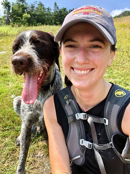 A smiling Kenney sits on the grass next to her dog.