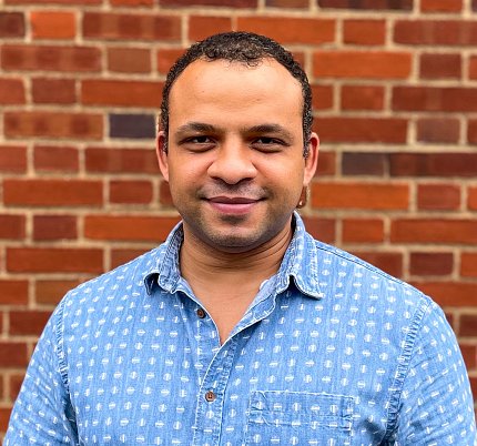 John Jima in a blue button-down shirt standing in front of a brick building