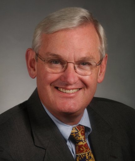 Henderson wears glasses and a black suit, with a blue shirt and gold, black, and red patterned tie. The photo background is gray.