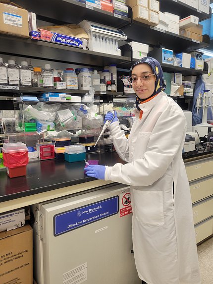 Fathi stands at a lab bench, holding a micropipette. Rows of shelving above the bench hold various lab materials.