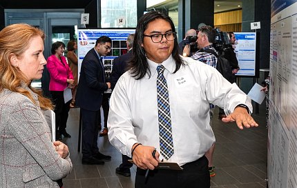 Woman stands viewing a poster as a young man in shirt & tie gestures toward it.