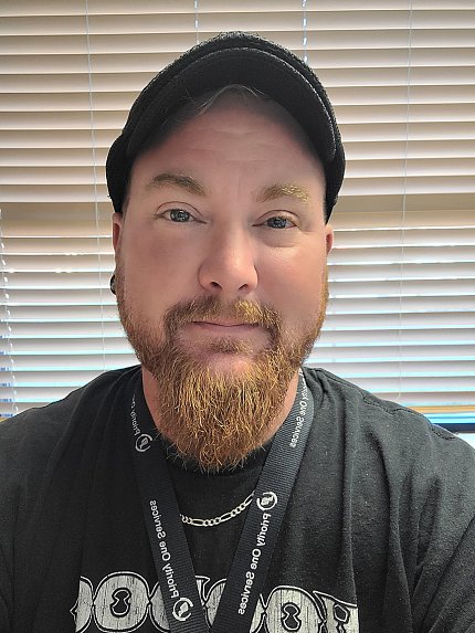 Harmon takes a selfie against a window with blinds. He is bearded and is wearing a dark baseball cap and black shirt.