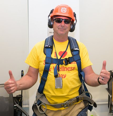 Man in bright yellow Wellness Day shirt, sunglasses, headphones, safety harness gives thumbs up.